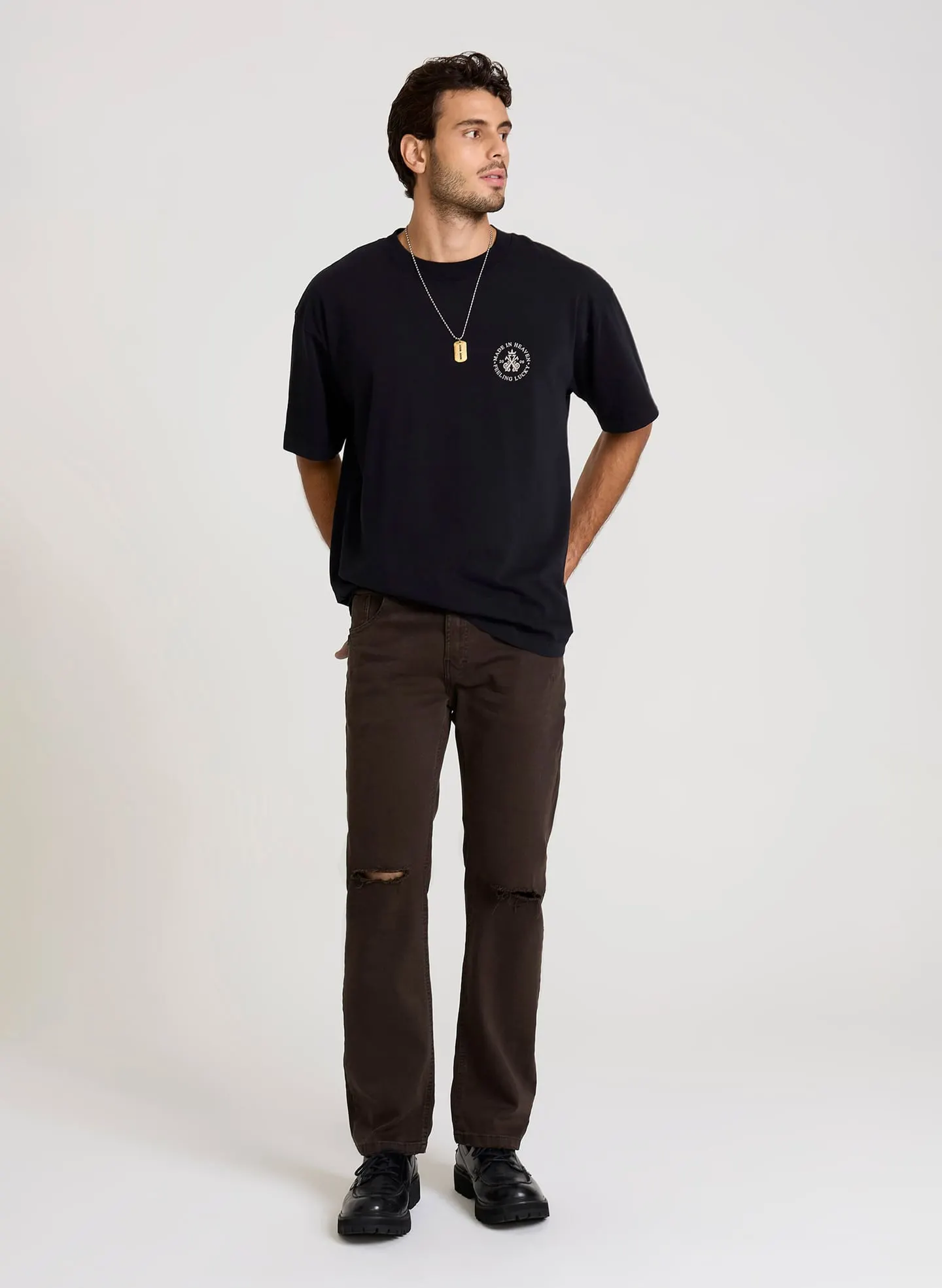 Homem vestindo camiseta preta de manga curta e calça marrom com rasgos nos joelhos